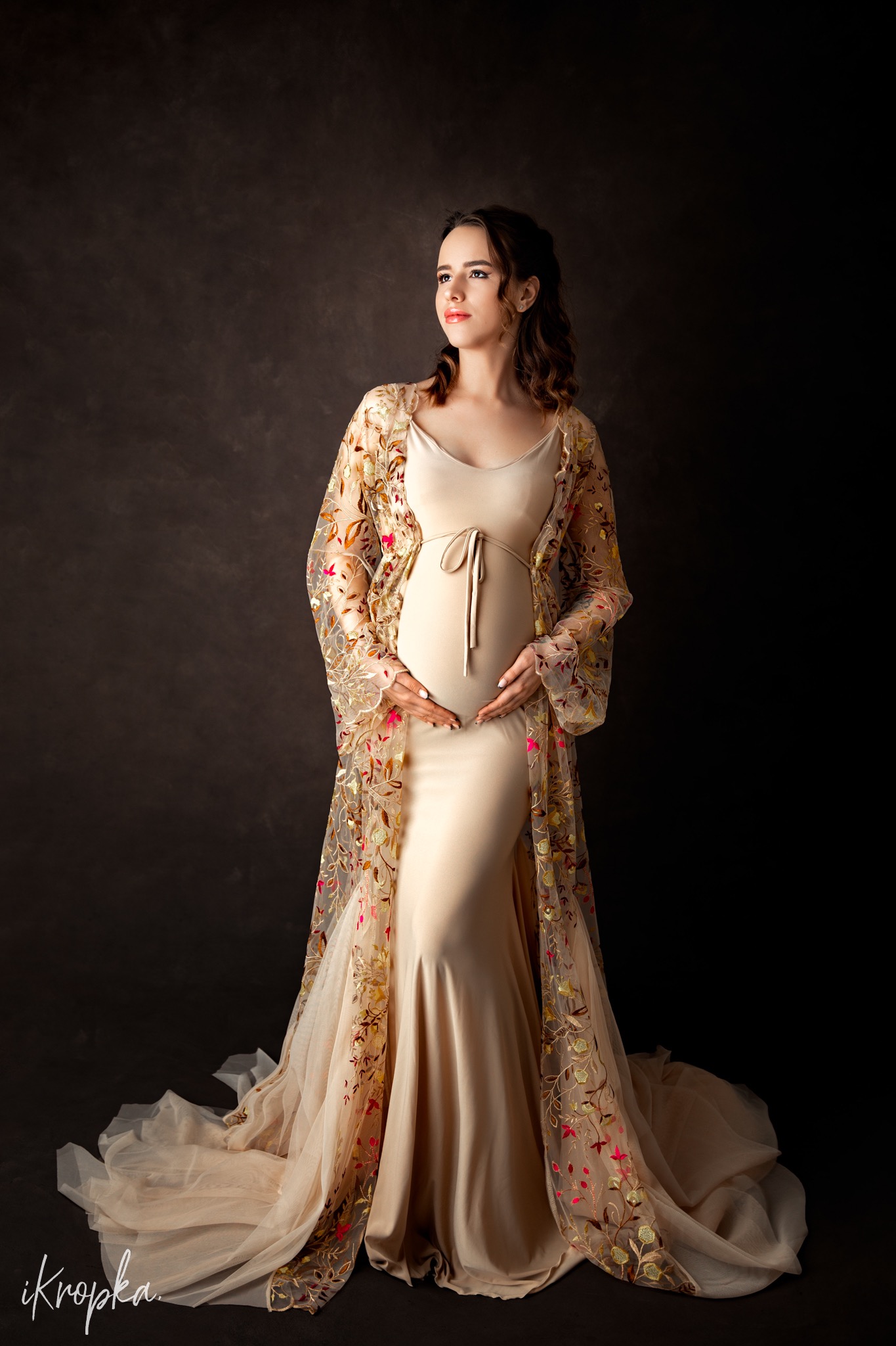 Kobieta w zaawansowanej ciąży, ubrana w beżową sukienkę i ozdobny, prześwitujący szlafrok z haftowanymi kwiatami, pozuje na ciemnym tle, trzymając dłonie na brzuchu i spoglądając w górę.