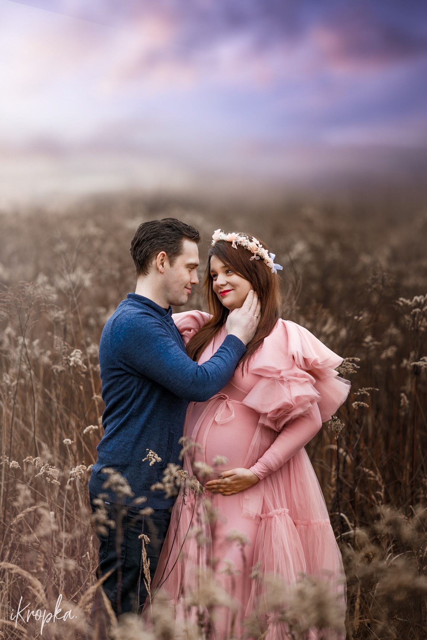 Przyszła mama w tiulowej sukni z falbanami i wiankiem na głowie, czule obejmowana przez mężczyznę na tle pola z uschniętą trawą i pastelowego nieba.