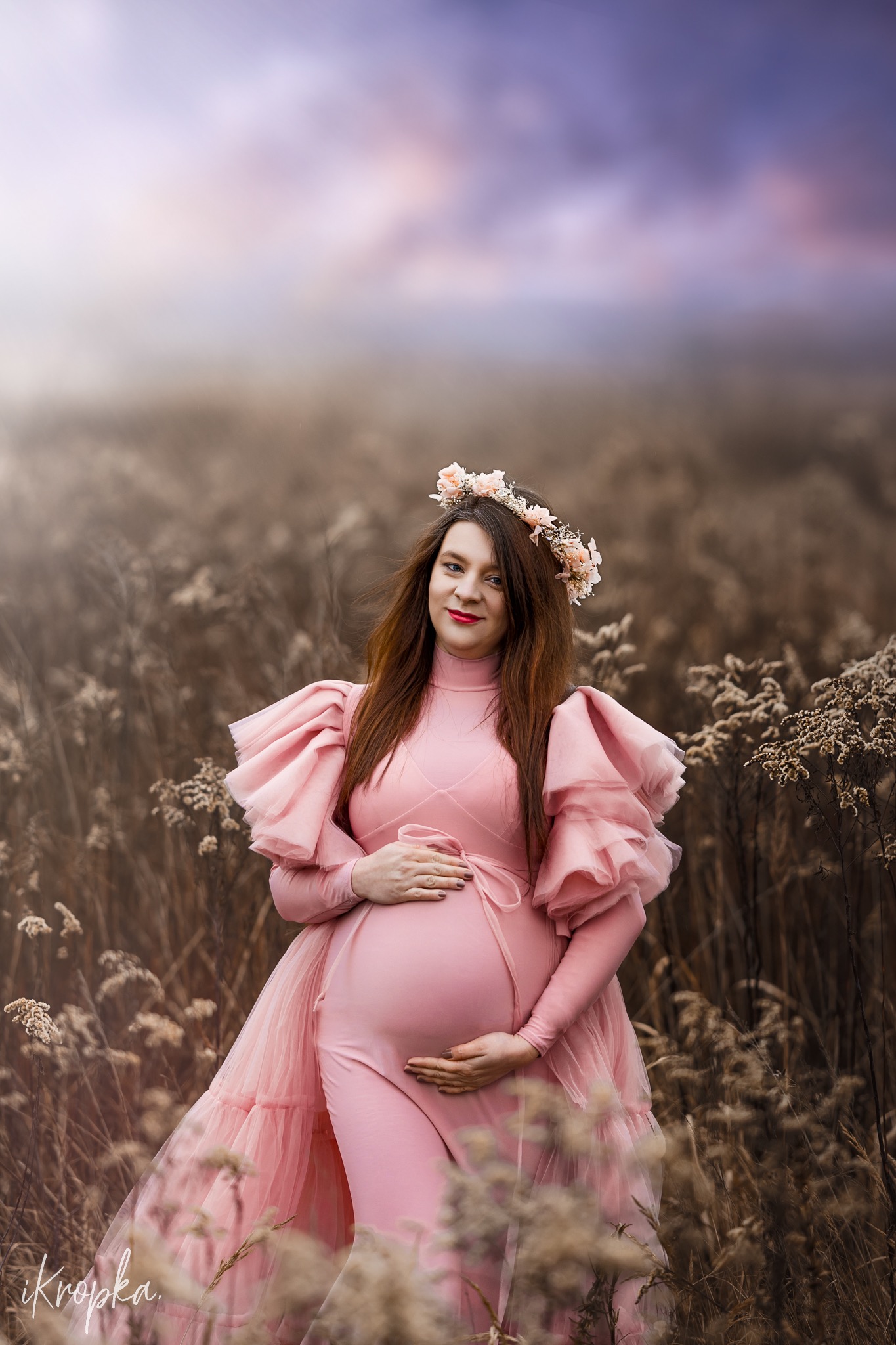 Kobieta w ciąży w różowej sukni z falbanami i kwiatowym wianku, pozująca na łące z uschniętą trawą, z rękami na brzuchu.