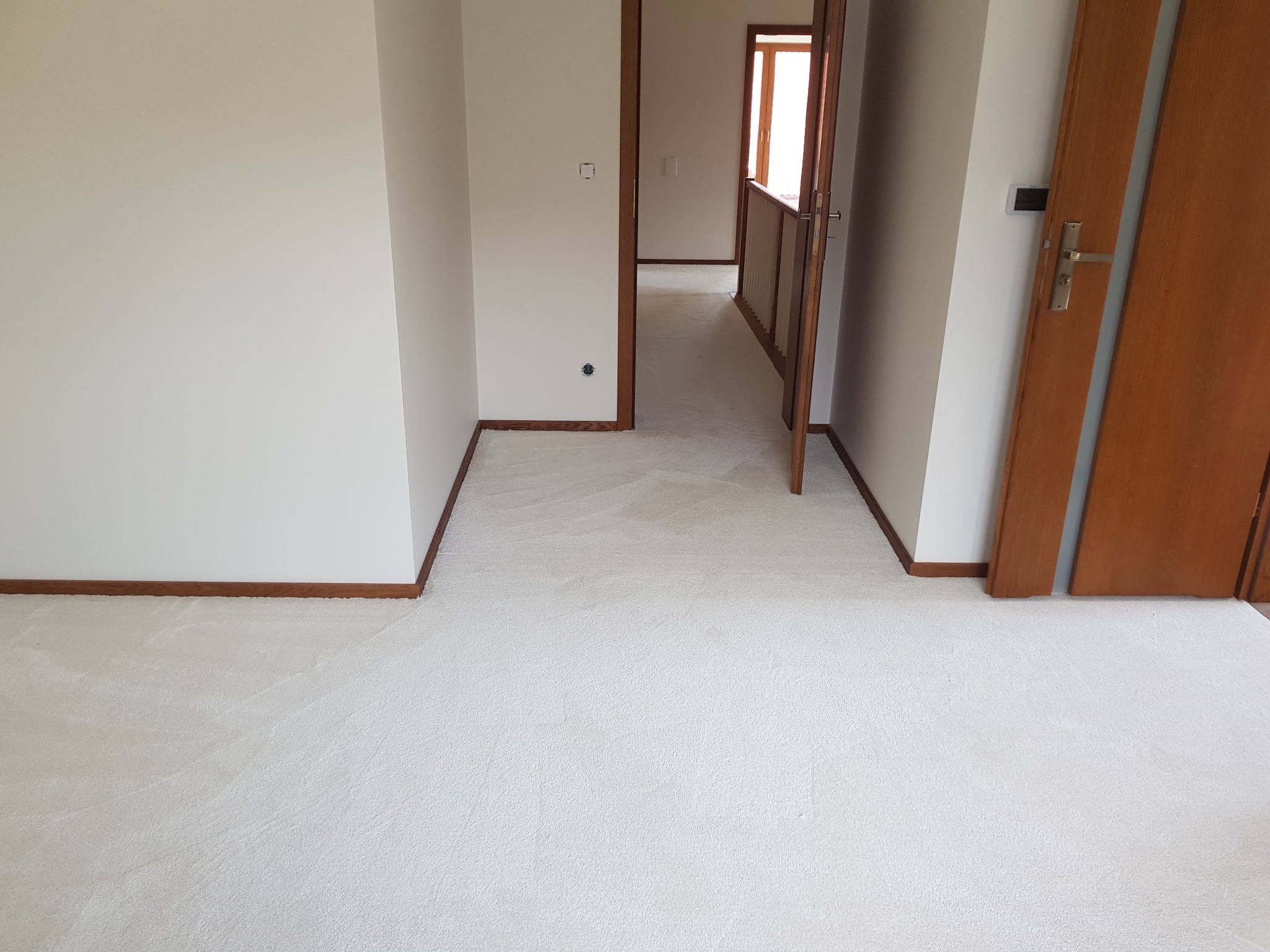 Jasna, kremowa wykładzina dywanowa w korytarzu z białymi ścianami i brązowymi listwami przypodłogowymi, widoczne otwarte drzwi z drewnianą ościeżnicą.