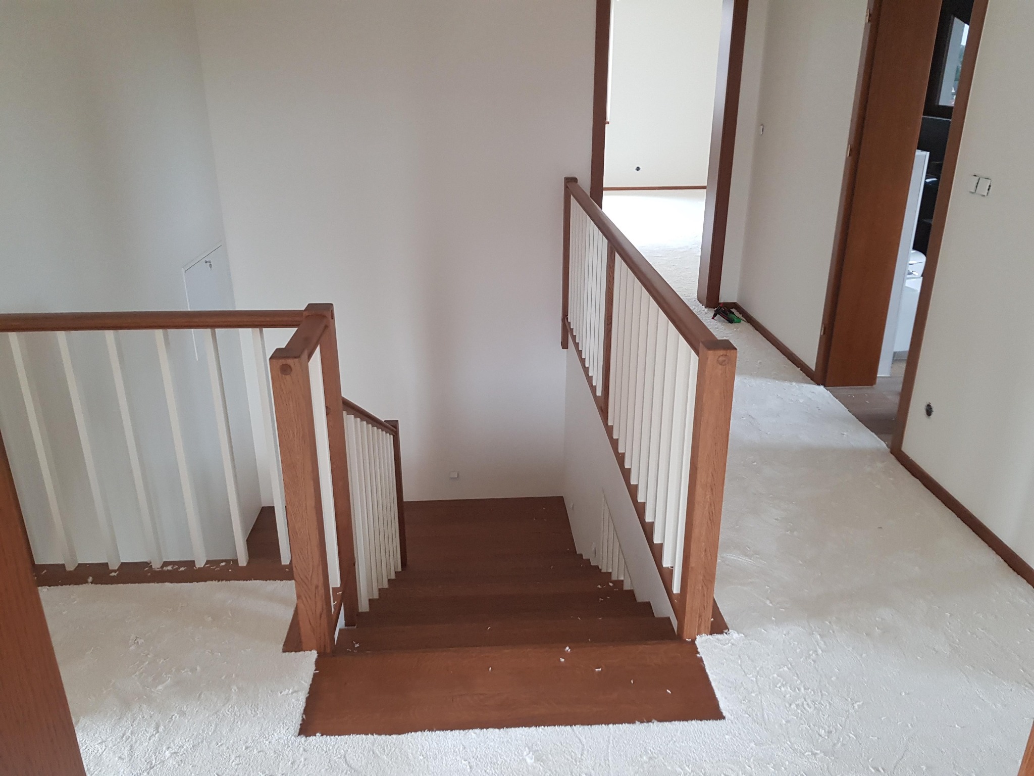 Widok z góry na drewniane schody z białymi balustradami w jasnym wnętrzu, z widocznymi śladami po remoncie na podłodze.