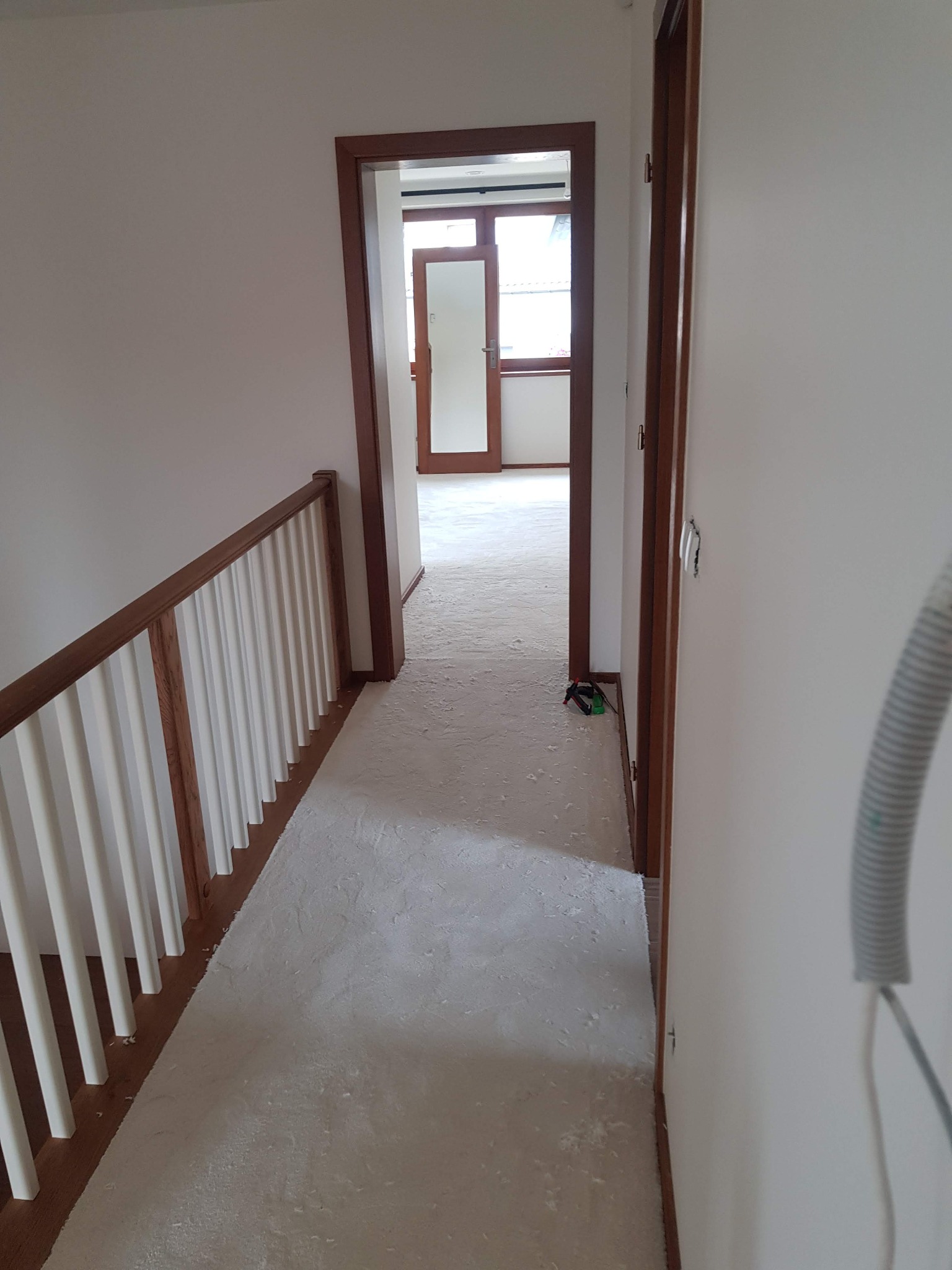 Korytarz w trakcie remontu z widoczną białą wykładziną pokrytą pyłem, drewnianą balustradą i ościeżnicami, narzędzia leżące na podłodze.