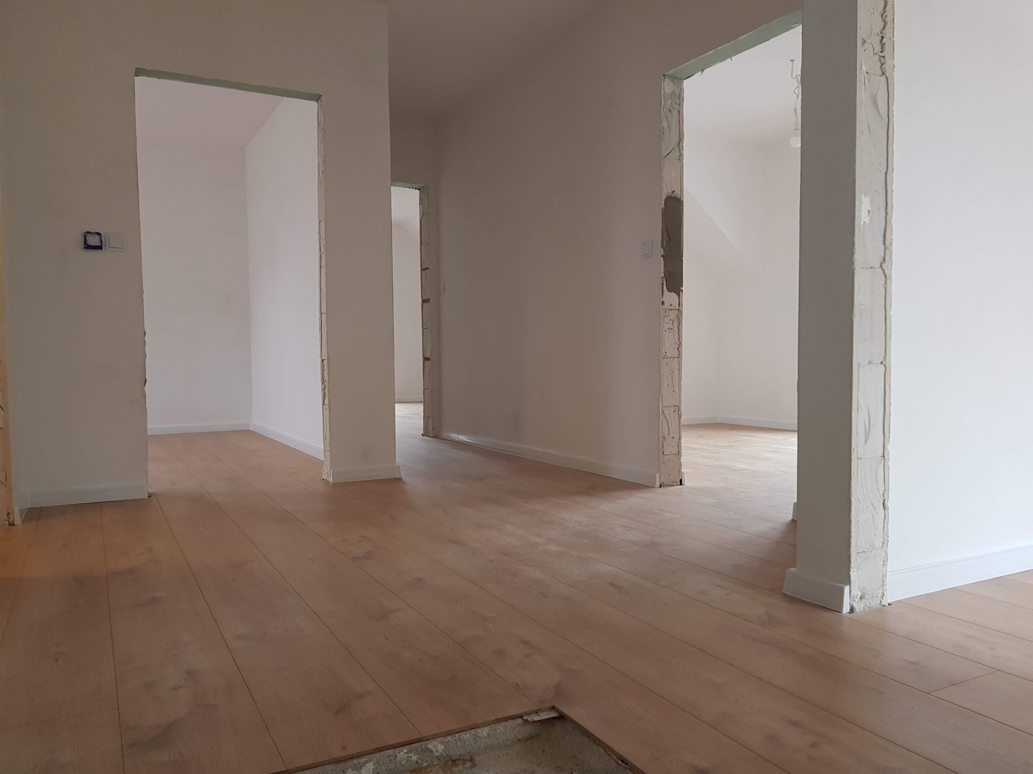 Jasne, drewniane deski podłogowe w trakcie montażu w pomieszczeniu z białymi ścianami i surowymi framugami drzwi, widoczny fragment nieukończonej podłogi z betonowym podłożem.