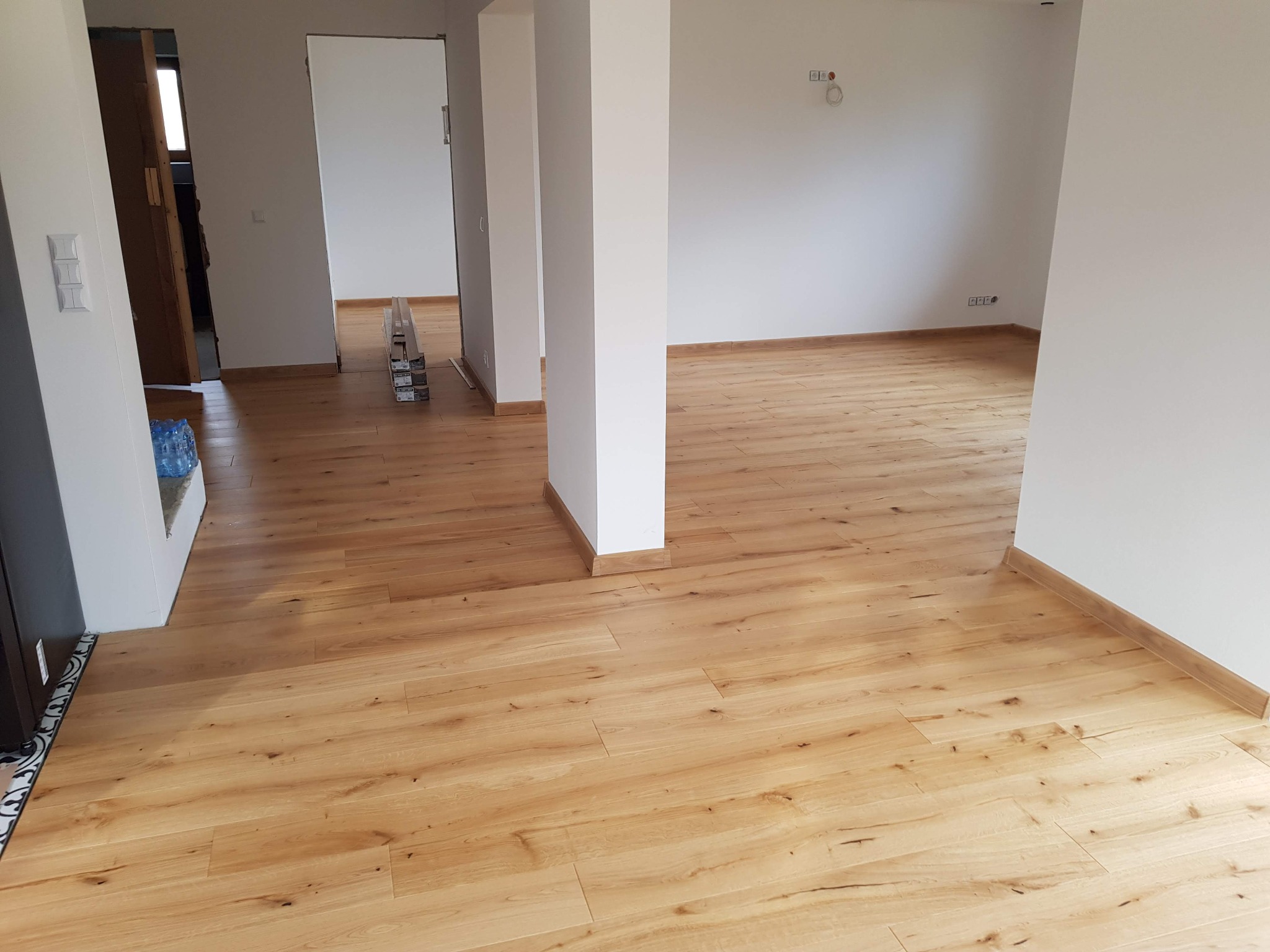 Jasna, nowa podłoga z paneli drewnopodobnych w przestronnym, jasnym wnętrzu. Widoczne białe ściany, kolumny i listwy przypodłogowe. W tle paczki paneli czekające na montaż.