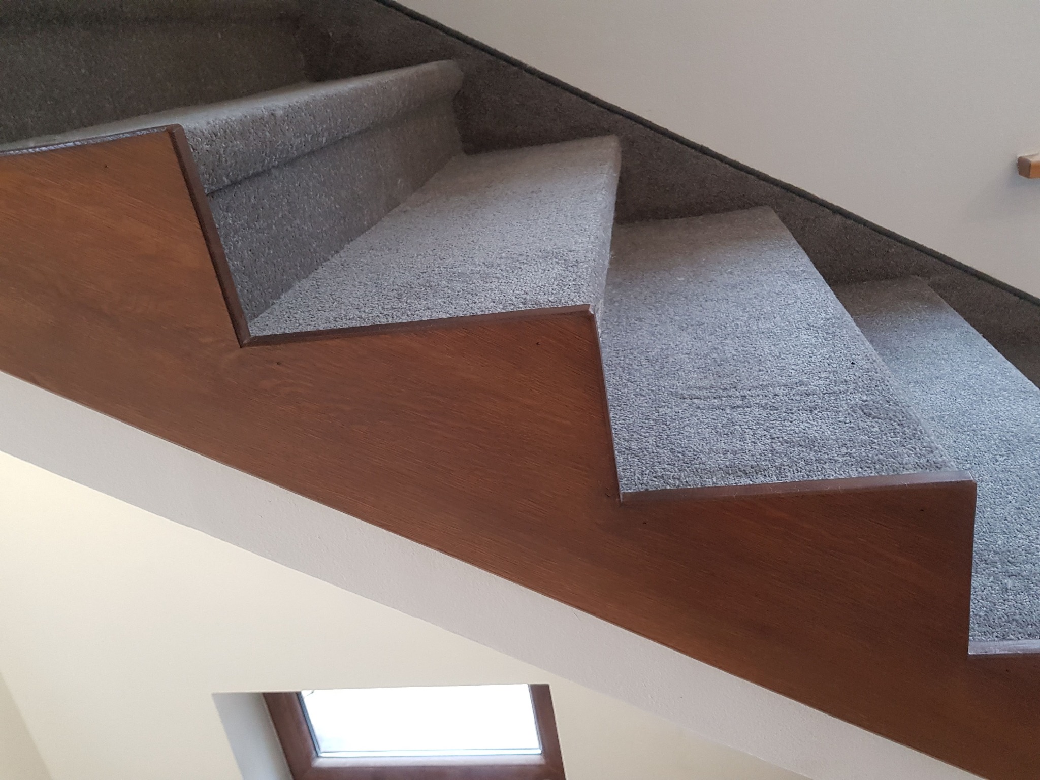 Schody z drewnianymi stopnicami i szarą wykładziną dywanową, widok z dołu, fragment okna w tle.