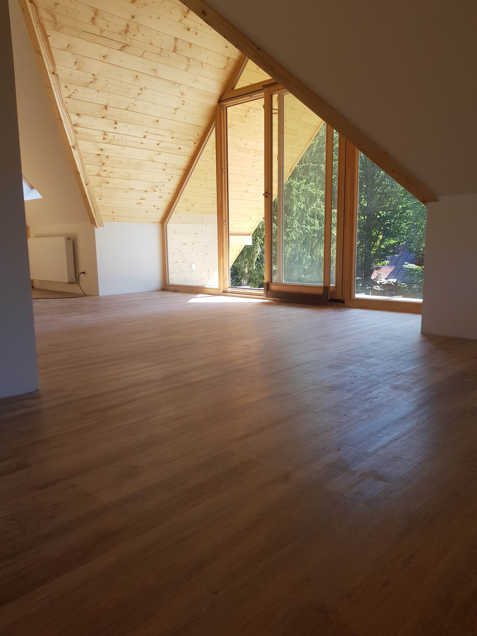 Jasne, drewniane poddasze z oknem szczytowym, świeżo położona podłoga z paneli, widok na zieleń za oknem.