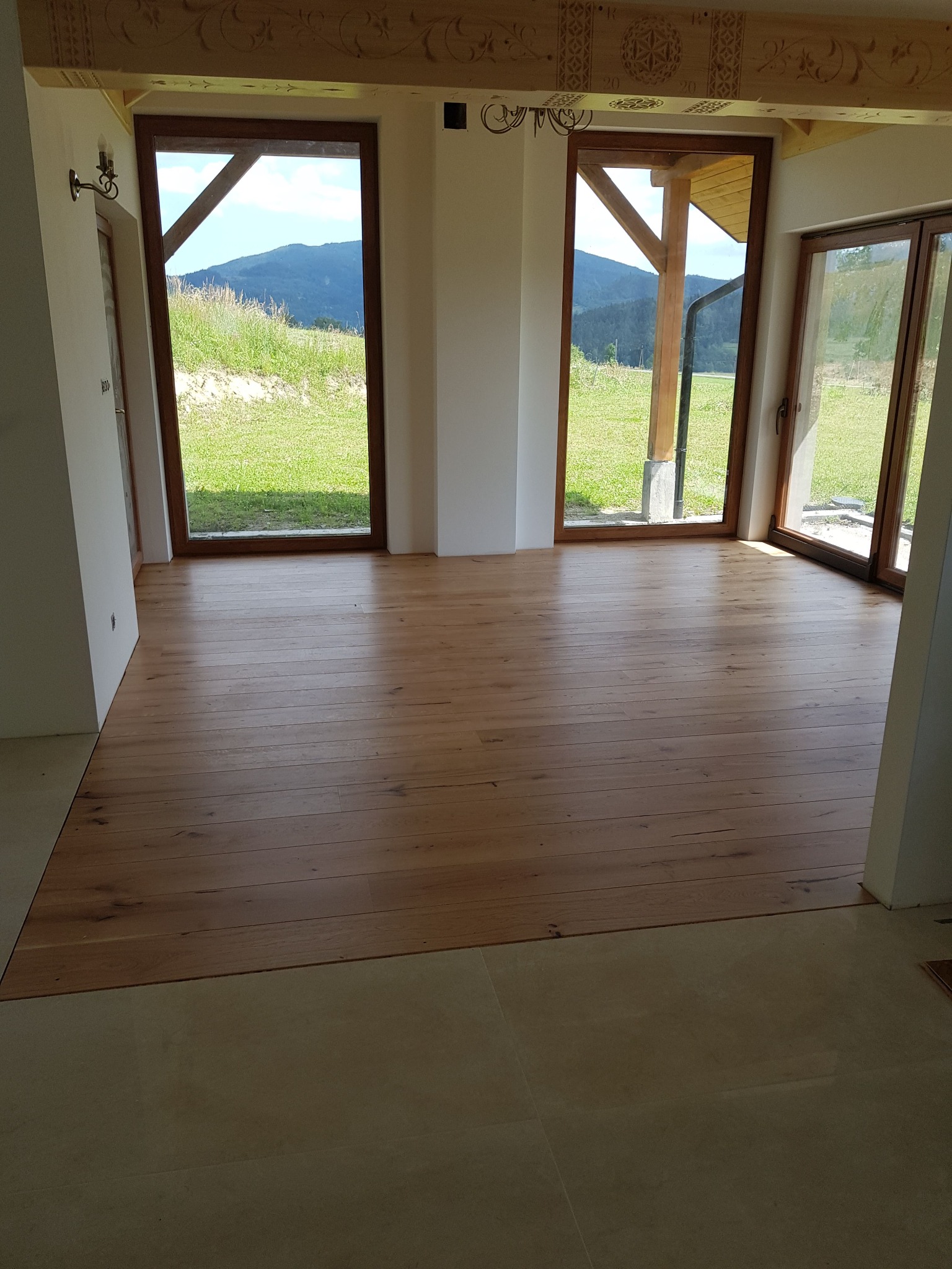 Jasny, drewniany parkiet w nowym domu z widokiem na góry przez duże okna; dekoracyjne drewniane belki sufitowe z rzeźbieniami.