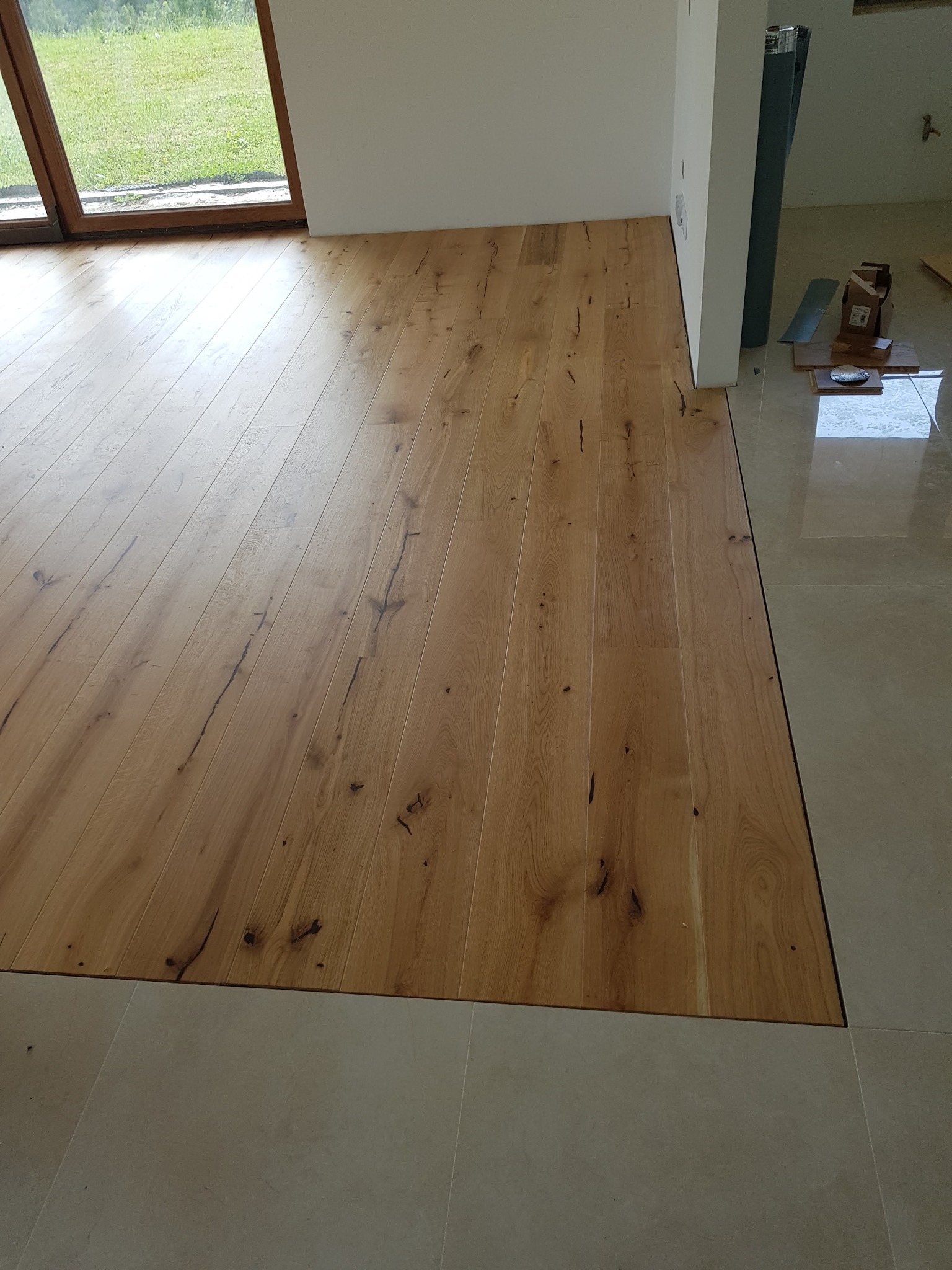 Jasne drewniane deski parkietu ułożone w nowym wnętrzu, widoczne przejście na podłogę z płytek, w tle okno z widokiem na zieleń.