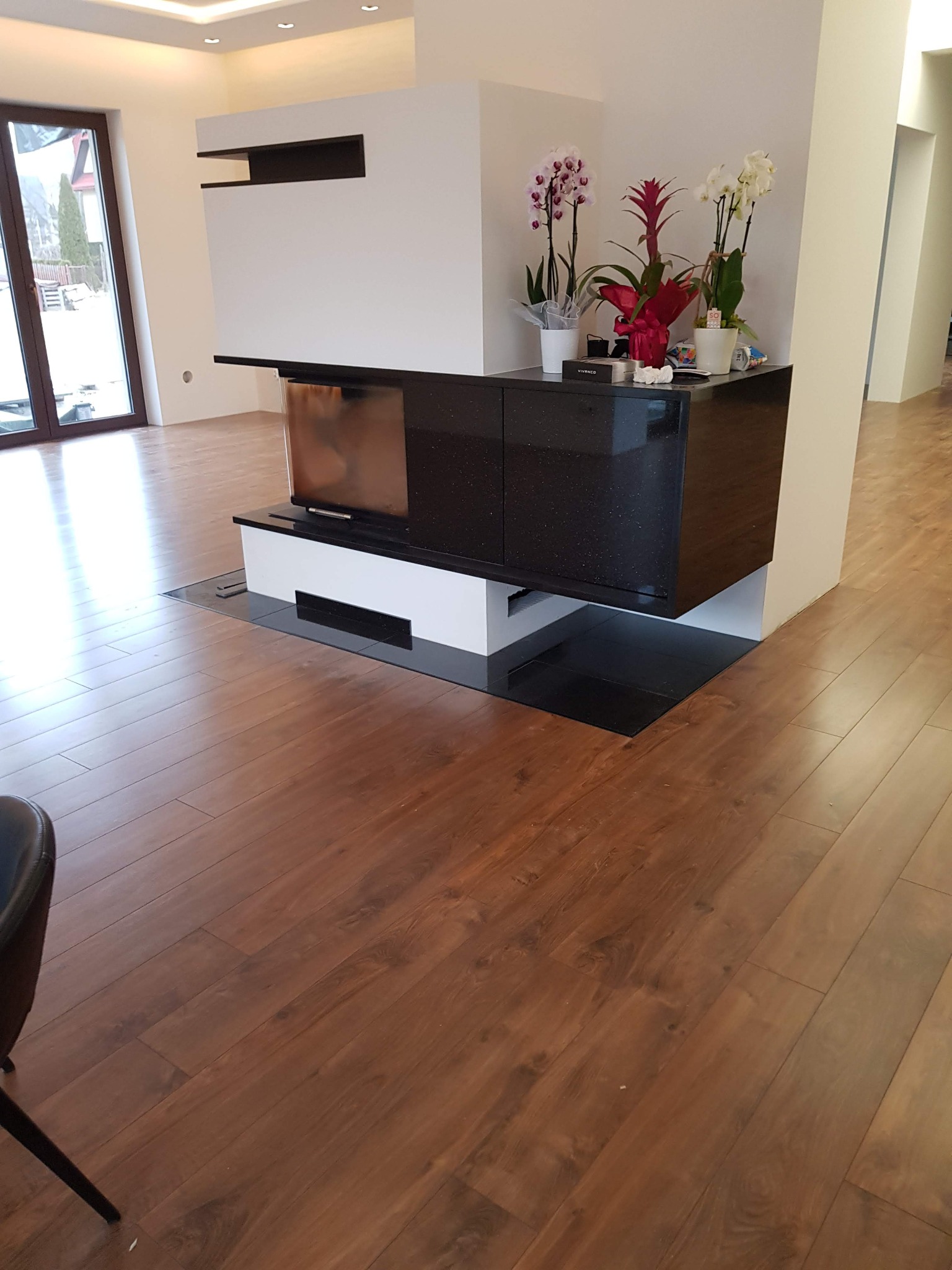 Ułożony parkiet w salonie z nowoczesnym kominkiem i meblami, widoczne deski parkietowe o ciepłym, brązowym odcieniu i wyraźnym usłojeniu, częściowo zasłonięte przez czarny mebel kominkowy z kwiatami.