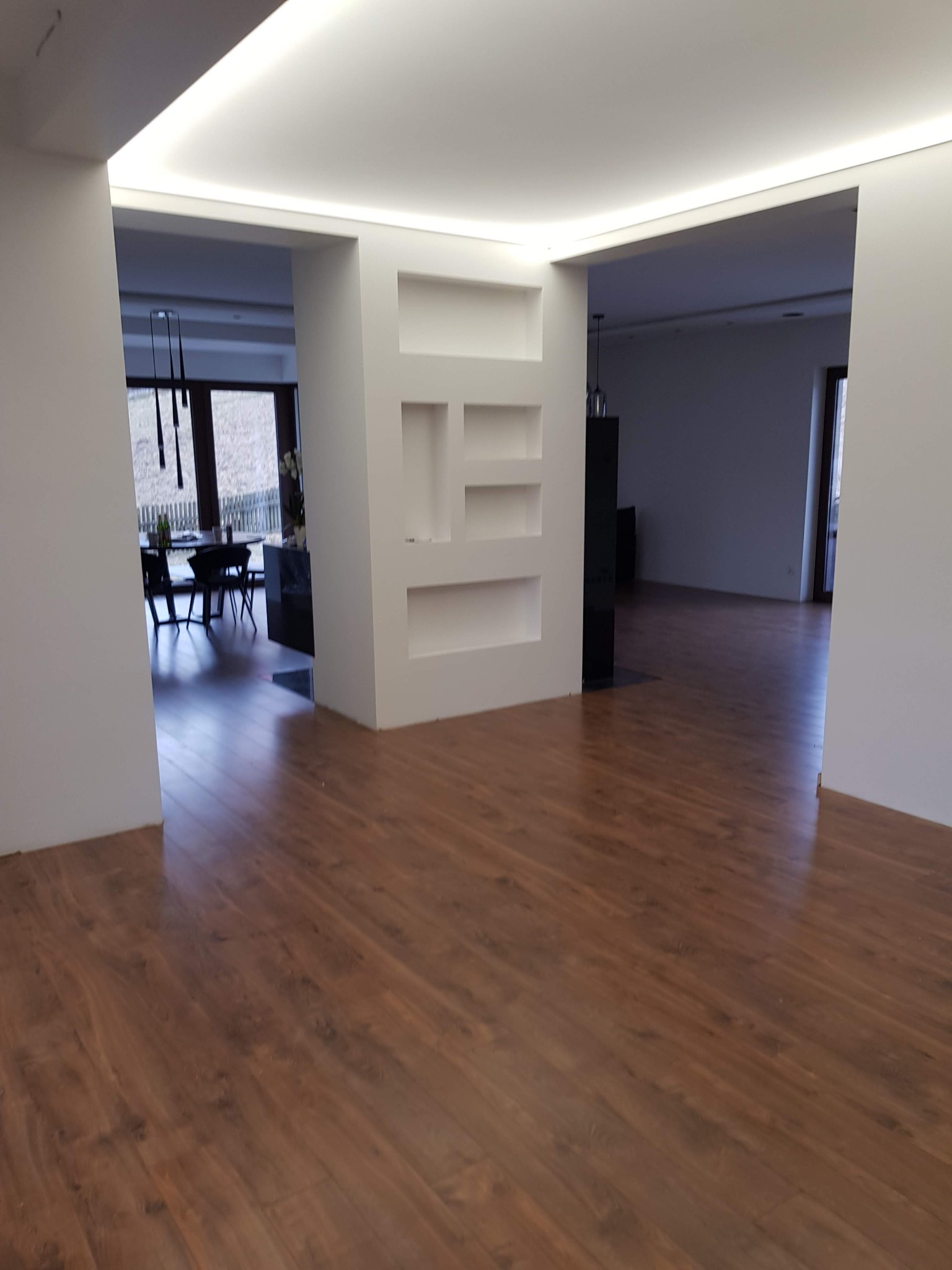 Ukończona podłoga z paneli w salonie z widokiem na jadalnię, oświetlenie LED w suficie, białe ściany z dekoracyjnymi wnękami.