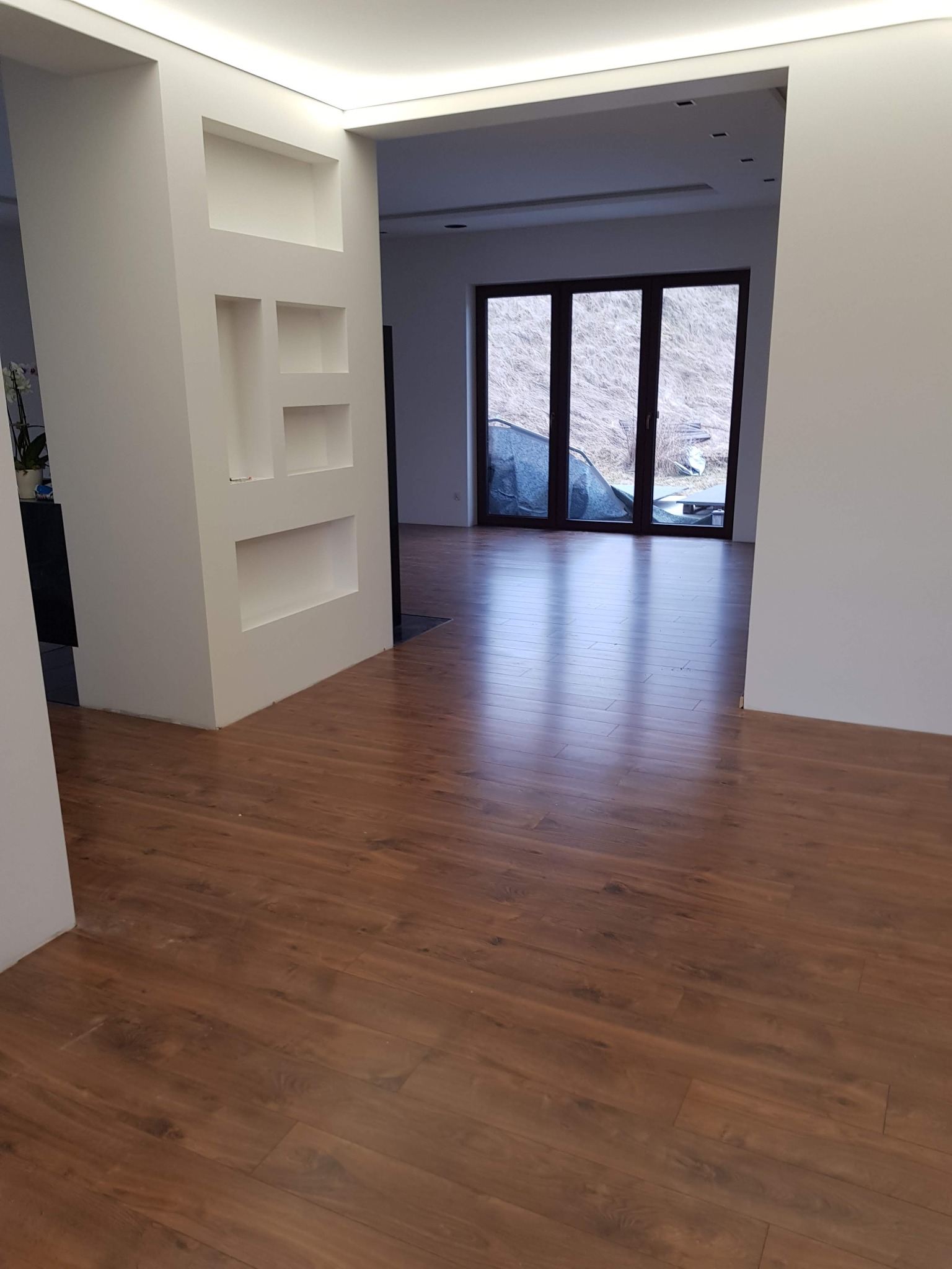 Nowo ułożona, błyszcząca podłoga z paneli w odcieniach brązu w przestronnym salonie w Nowym Targu, z widocznymi białymi ścianami z wnękami i dużym oknem tarasowym z widokiem na zewnątrz.