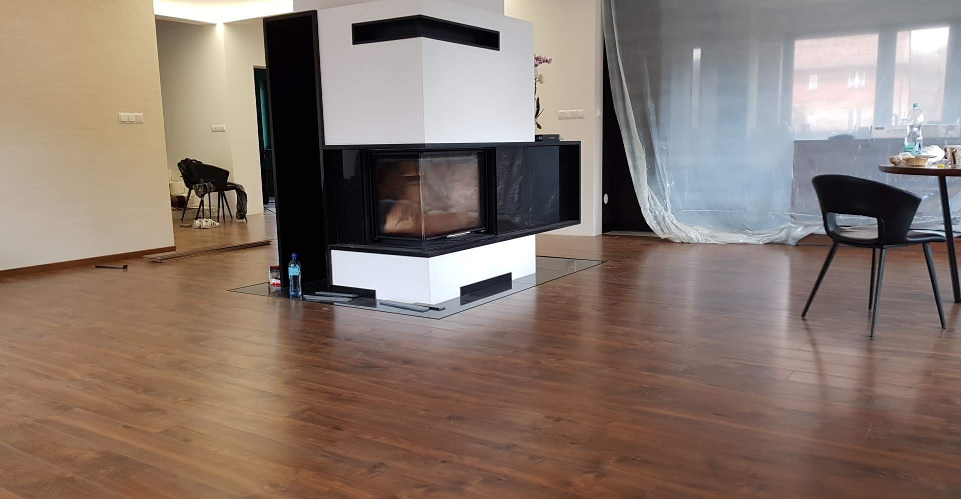 Ułożona podłoga z paneli w odcieniach brązu, widoczny fragment kominka z szybą, zabezpieczone folią okno, butelka wody i niedokończone listwy przypodłogowe.