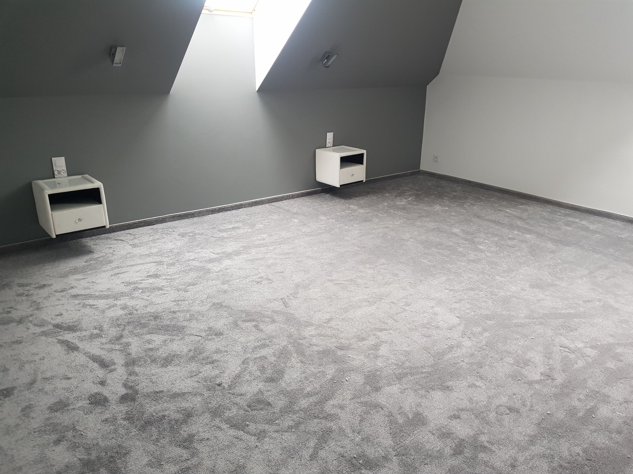 Szara, wzorzysta wykładzina w pokoju na poddaszu z oknem dachowym i dwiema białymi szafkami nocnymi.