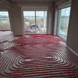 Czerwone rury ogrzewania podłogowego ułożone na folii w surowym wnętrzu z widokiem na zewnątrz przez okna.