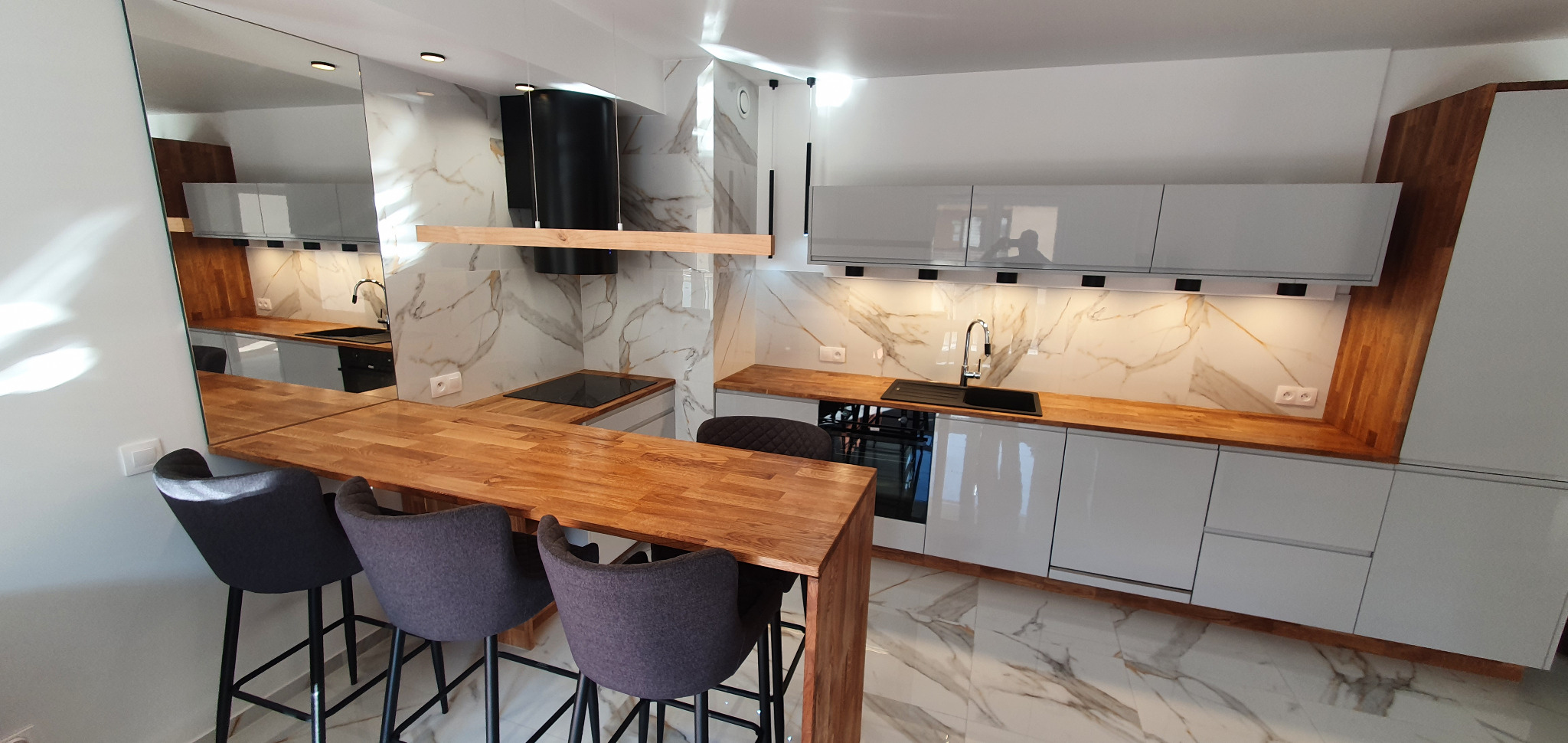 Nowoczesna kuchnia z białymi szafkami, drewnianym blatem i marmurową ścianą, uzupełniona o czarne elementy takie jak okap i zlew oraz wysokie krzesła barowe.