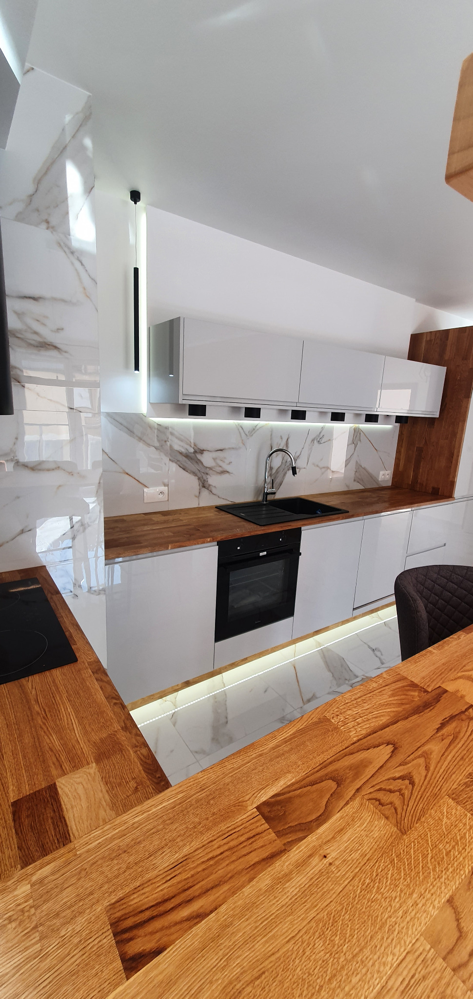 Nowoczesna kuchnia z białymi, lakierowanymi szafkami, blatem z drewna i marmurowymi płytkami na ścianie, oświetlona listwą LED pod szafkami i wiszącą lampą.