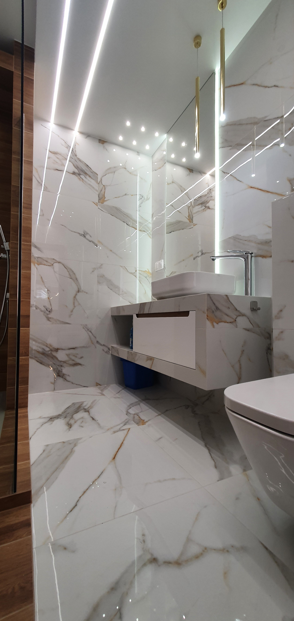 Nowoczesna łazienka z białymi płytkami imitującymi marmur, podświetlanym lustrem, wiszącą szafką z umywalką i złotymi lampami wiszącymi.