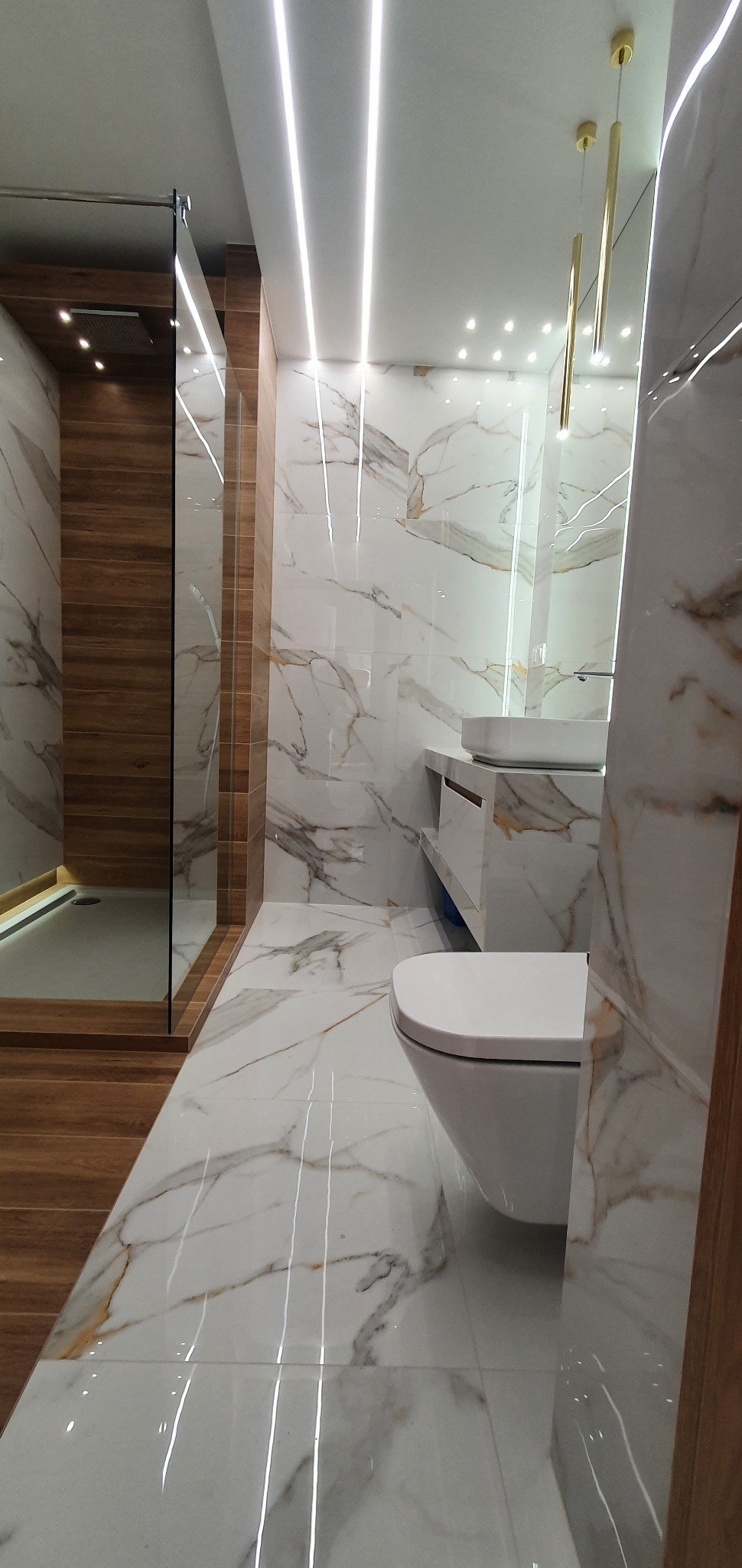 Nowoczesna łazienka z marmurowymi płytkami, oświetleniem LED w suficie i złotymi lampami wiszącymi. Kabina prysznicowa z drewnianymi akcentami.