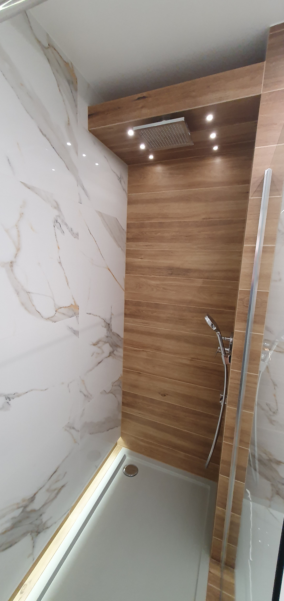 Nowoczesna kabina prysznicowa walk-in z drewnopodobną ścianą, deszczownicą sufitową z oświetleniem punktowym i marmurowymi płytkami na ścianach.