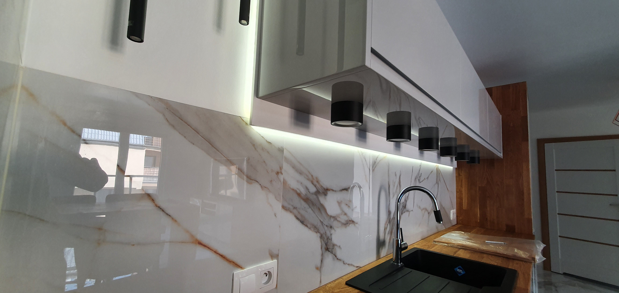 Nowoczesna kuchnia z białymi szafkami, marmurową ścianą, czarnym zlewem i oświetleniem punktowym podszafkowym.