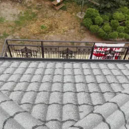 Widok z góry na dach pokryty szarym gontem bitumicznym, z ozdobną metalową balustradą balkonu w tle i fragmentem ogrodu.