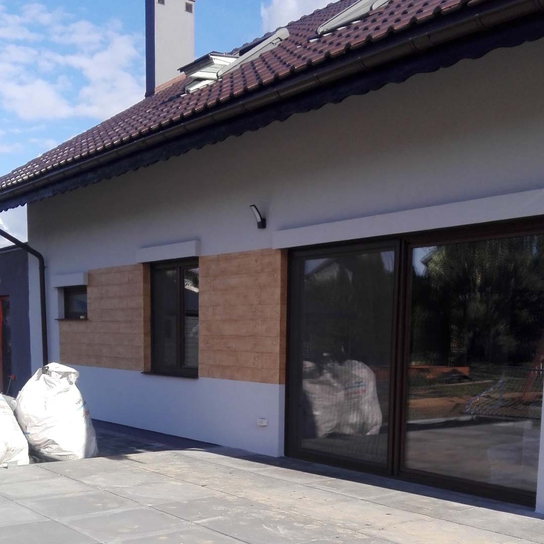 Elewacja domu jednorodzinnego z fragmentem ściany wykończonym imitacją cegły, widoczne okna z ciemnymi ramami i duże przesuwne drzwi tarasowe, na pierwszym planie worki z materiałami budowlanymi.