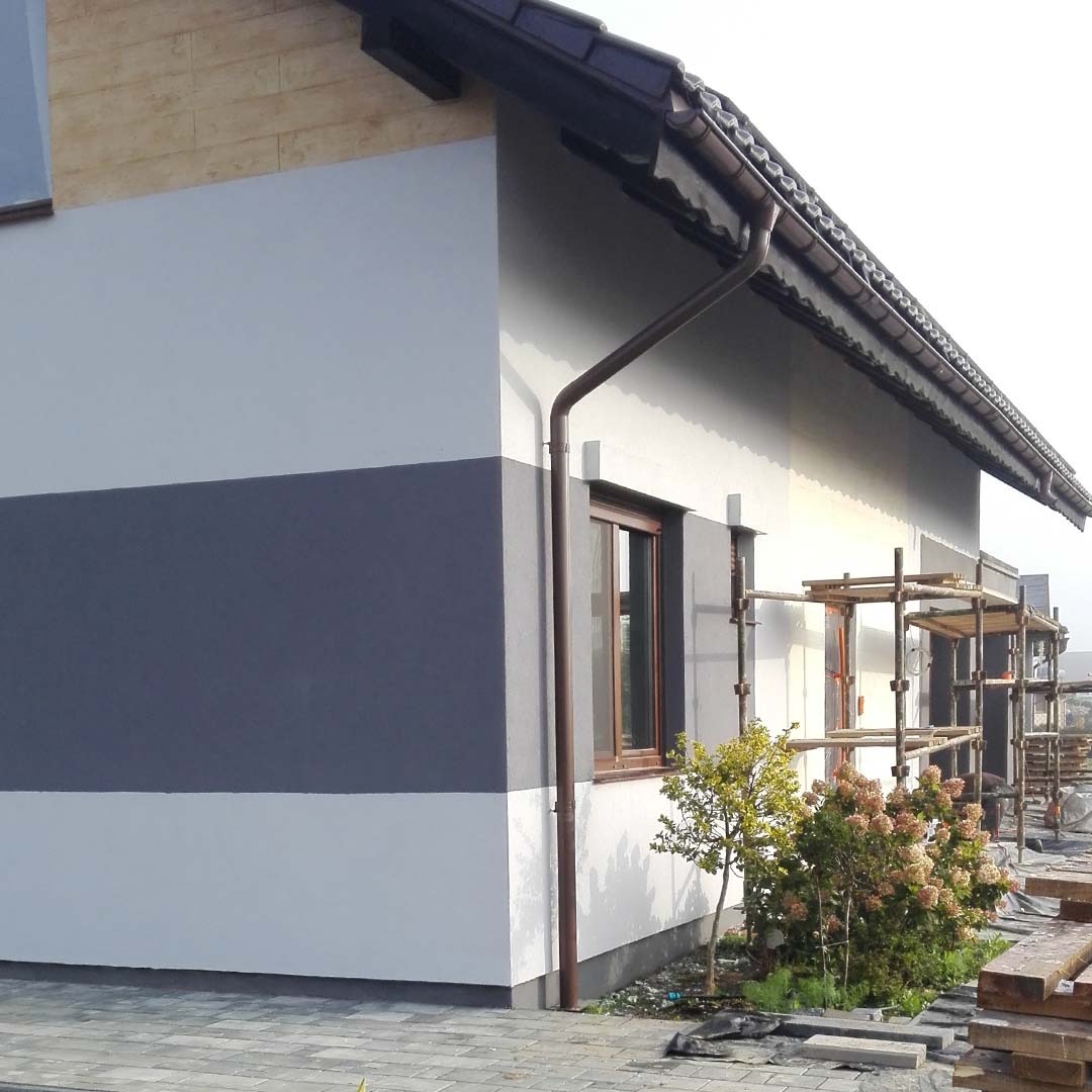 Dwukolorowa elewacja domu jednorodzinnego, z widocznymi rusztowaniami i elementami drewnianymi na górze ściany, okno z brązową ramą i rynna w kolorze brązowym, kostka brukowa przed budynkiem.