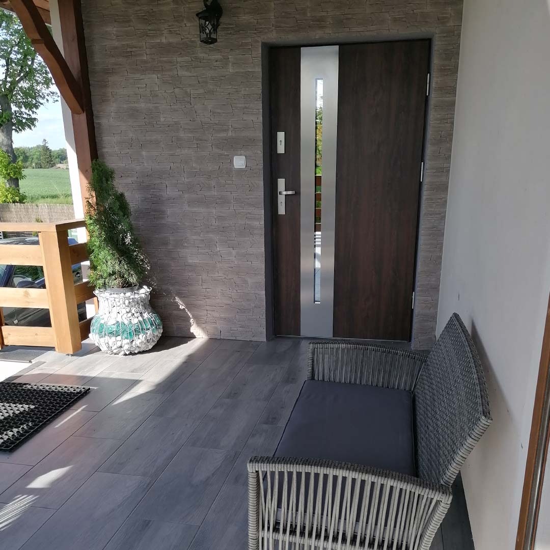 Zadaszone wejście do domu z grafitową podłogą imitującą drewno, kamienną elewacją, ciemnymi drzwiami z pionowym przeszkleniem, dekoracyjną donicą z iglakiem i rattanową ławką z szarą poduszką.