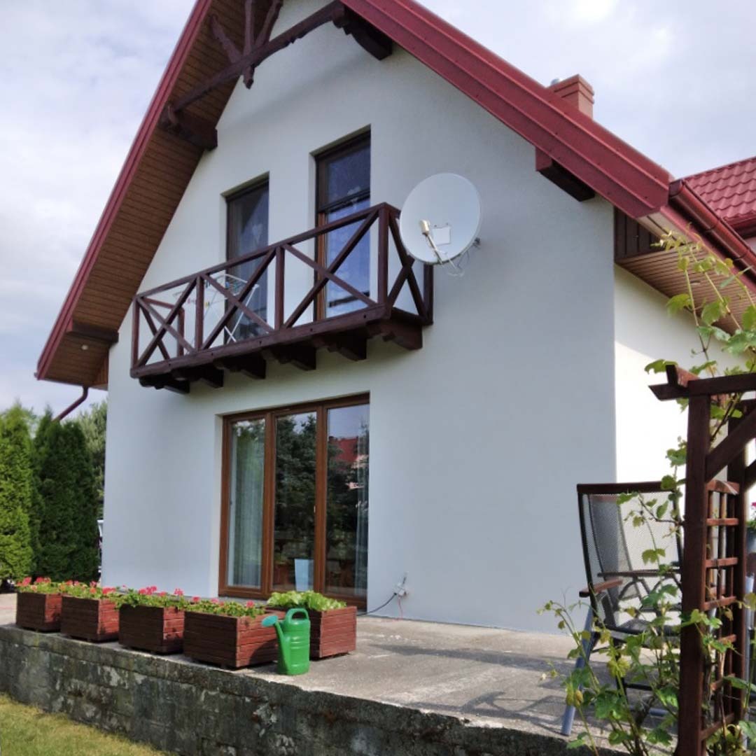 Biały dom jednorodzinny z czerwonym dachem i drewnianym balkonem, widoczna antena satelitarna, kwiaty w donicach i zielony konewka na tarasie.