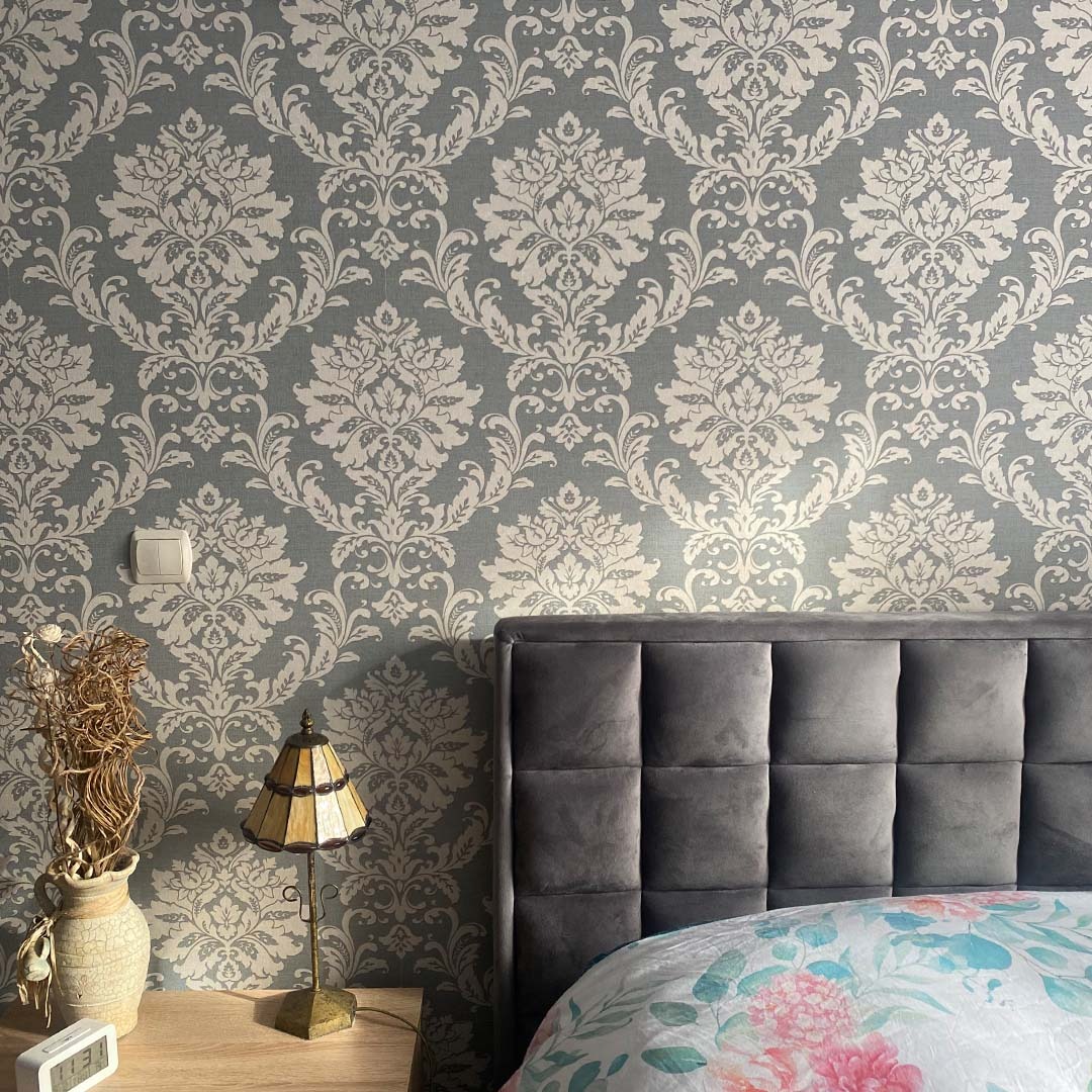 Szara ściana w ozdobny, biały wzór damaskowy w sypialni, widoczny fragment łóżka z szarym, welurowym zagłówkiem i pościelą w kwiaty, na szafce nocnej lampka w stylu retro i wazon z suchymi gałązkami.