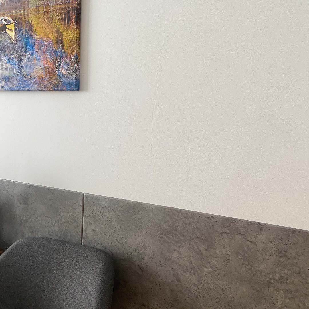Biała ściana z obrazem przedstawiającym łodzie i abstrakcyjny krajobraz, poniżej szara, kamienna okładzina ścienna i fragment szarego krzesła