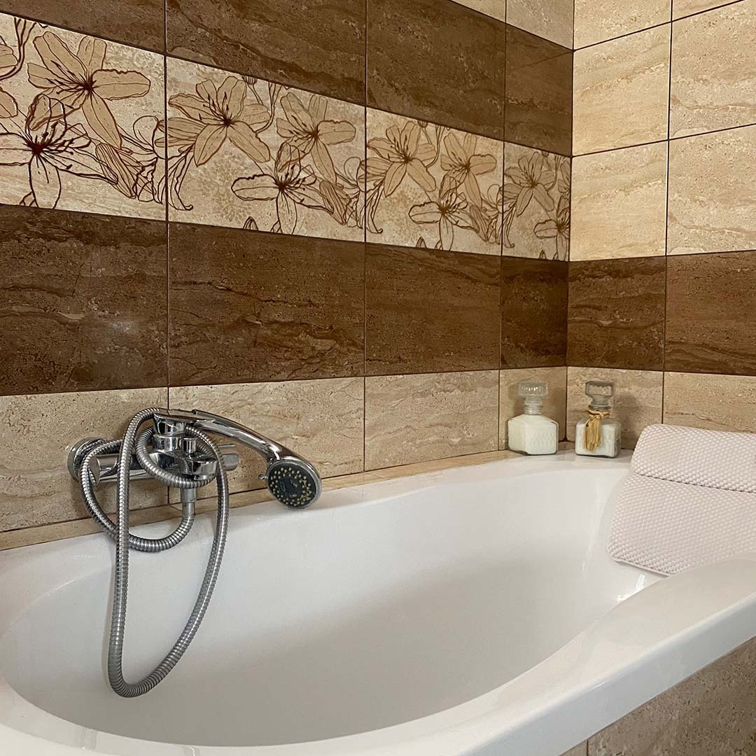 Biała wanna z chromowaną baterią i słuchawką prysznicową na tle ściany wyłożonej brązowo-beżowymi płytkami z motywem kwiatowym, uzupełniona dekoracyjnymi flakonami i poduszką.