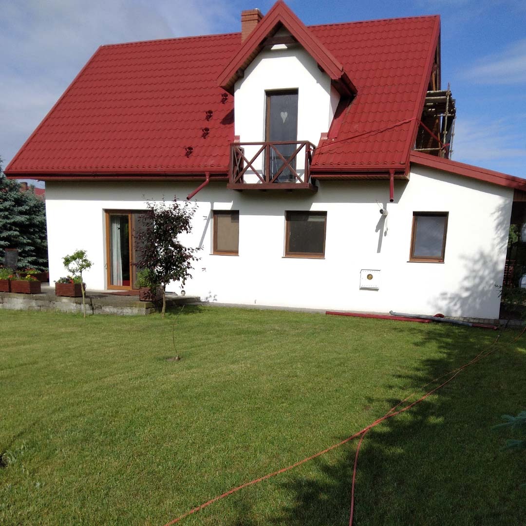 Biały dom jednorodzinny z czerwoną dachówką, drewnianym balkonem i widocznym rusztowaniem z boku, otoczony zielonym trawnikiem. Widoczne trzy okna z brązowymi ramami oraz drzwi balkonowe.