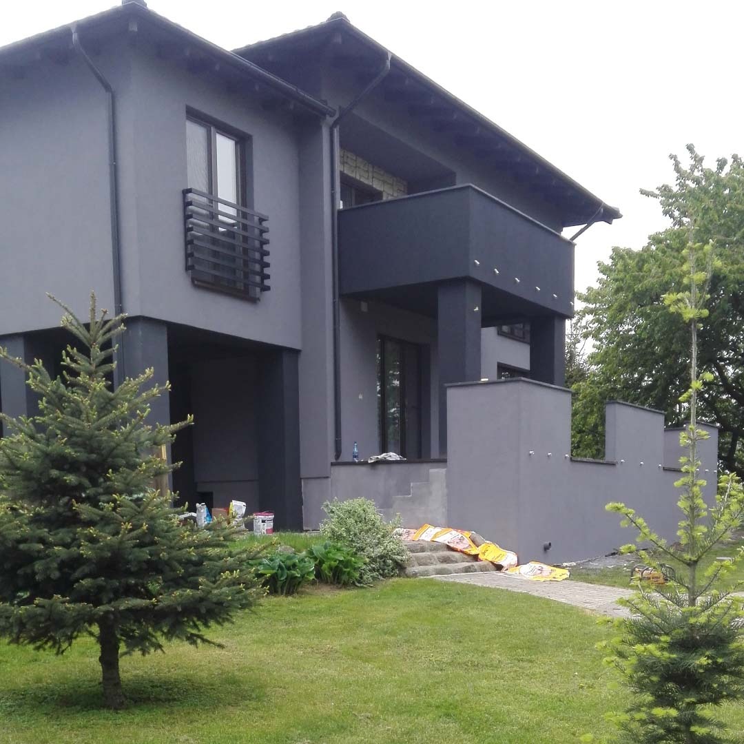 Nowoczesny dom z szarą elewacją, widoczne puszki z farbą i worki z materiałem na schodach wejściowych, trawnik z młodymi drzewkami iglastymi.