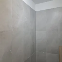 Ściana wyłożona szarymi, matowymi płytkami imitującymi marmur, z widocznym czarnym przyciskiem spłuczki i fragmentem drewnianych drzwi.