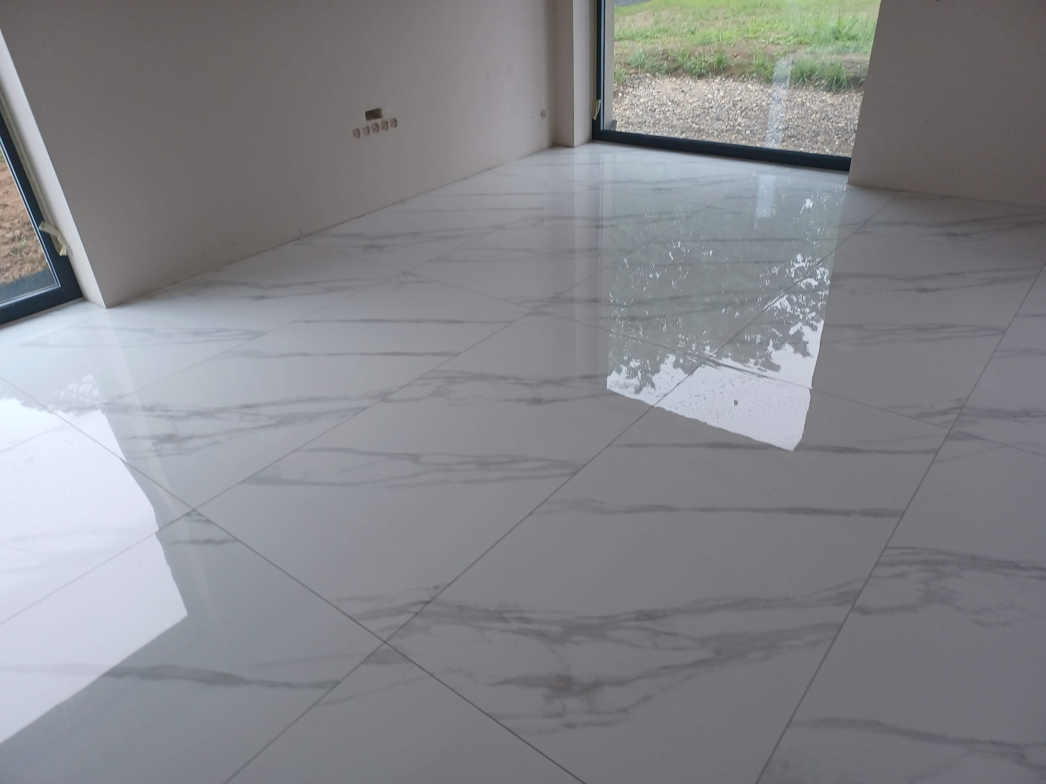 Błyszcząca, biała podłoga z płytek imitujących marmur w nowym, jasnym pomieszczeniu z dużymi oknami i widocznymi gniazdkami elektrycznymi na ścianie.