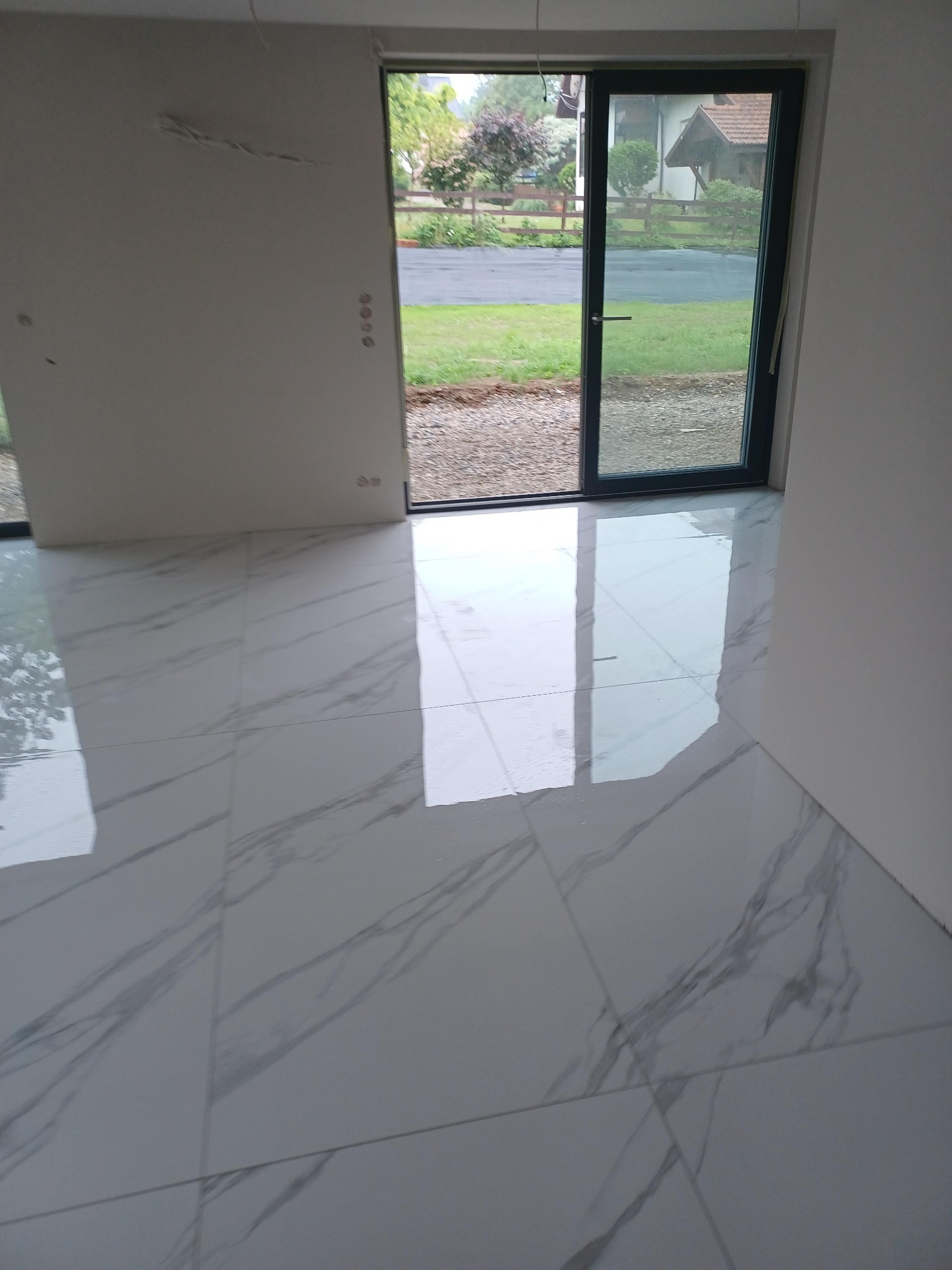 Błyszcząca, jasnoszara podłoga z płytek imitujących marmur, świeżo położona w pustym pomieszczeniu z widokiem na ogród przez otwarte drzwi balkonowe.