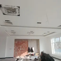 Wnętrze w trakcie wykańczania: fragment ściany z cegły, częściowo otynkowana ściana, sufit z podwieszanym oświetleniem w folii ochronnej, widoczne kable, sprzęt budowlany przykryty folią.