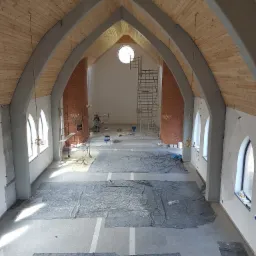 Wnętrze kościoła w trakcie remontu, widoczne rusztowania, zabezpieczona podłoga folią, drewniana boazeria na sklepieniu i pracownik malujący ścianę.