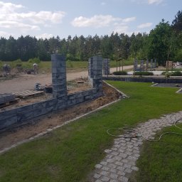 Usługi remontowe - Budowa ogrodzenia z szarych bloczków betonowych, widoczny fragment trawnika, ścieżka z kostki brukowej i pracownik z taczką w tle.