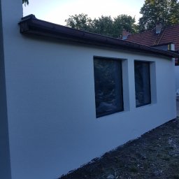 Usługi remontowe - Świeżo pomalowana, biała ściana budynku z dwoma oknami, widok z zewnątrz, nierówny teren u podstawy ściany, w tle drzewa i dach sąsiedniego budynku.