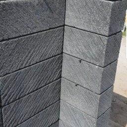 Usługi remontowe - Wykończony narożnik elewacji z ciemnoszarych, teksturowanych płyt kamiennych ułożonych w regularny wzór, widoczny fragment trawnika i osoba w tle.