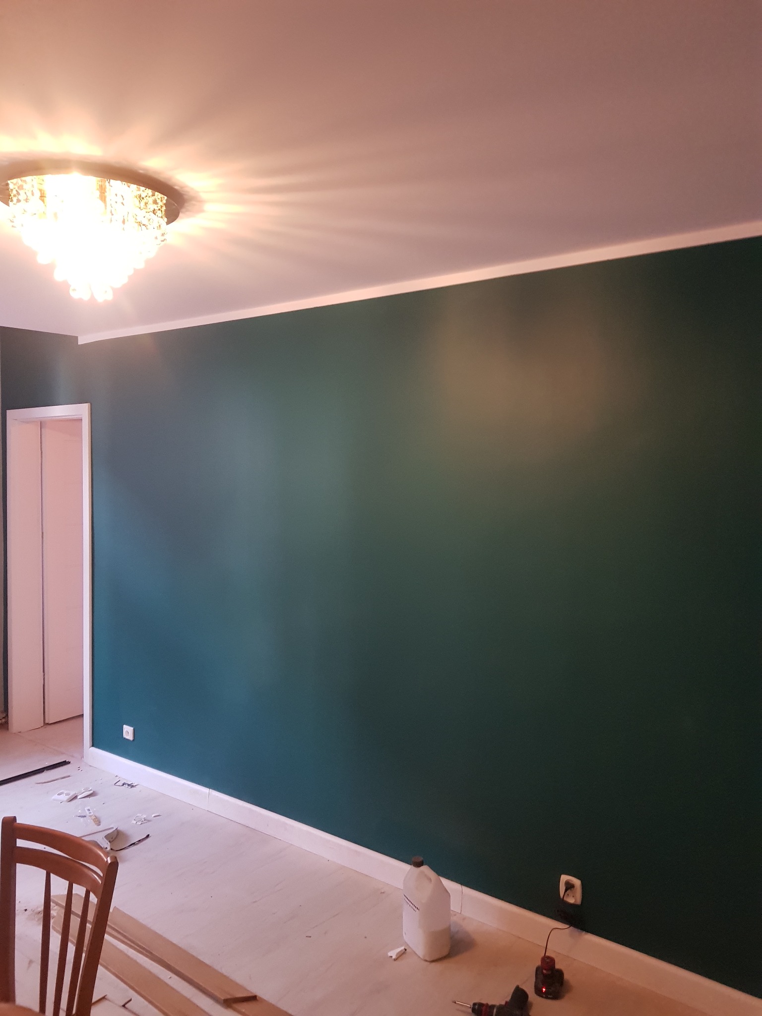 Świeżo pomalowana ściana w odcieniach zieleni, z widocznym kryształowym żyrandolem, listwami przypodłogowymi w trakcie montażu, narzędziami i puszką farby na podłodze.