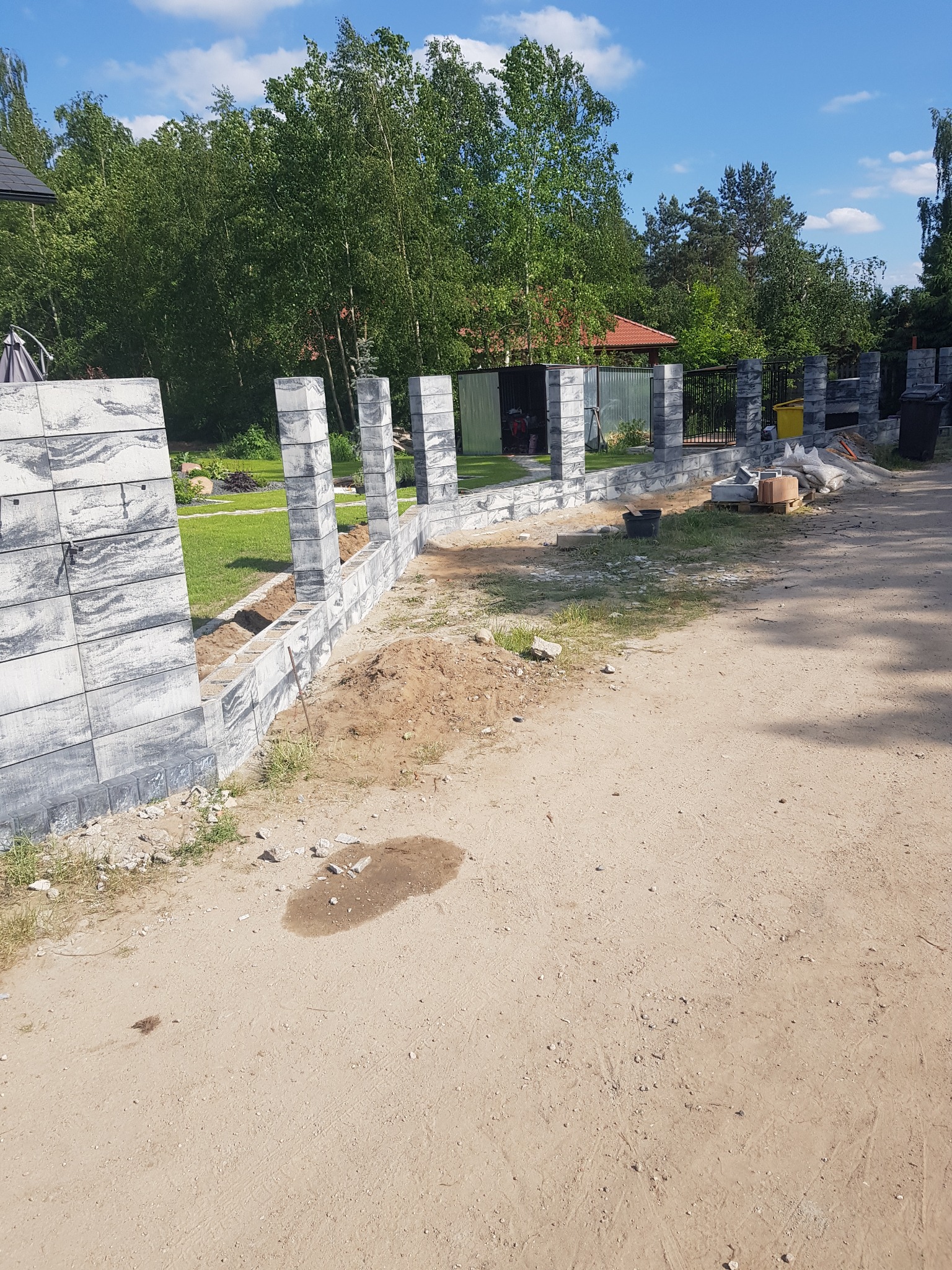 Budowa ogrodzenia z prefabrykatów betonowych o fakturze drewna, widoczne słupki i fragmenty muru w trakcie montażu, teren budowy z piaskiem i zielenią w tle.