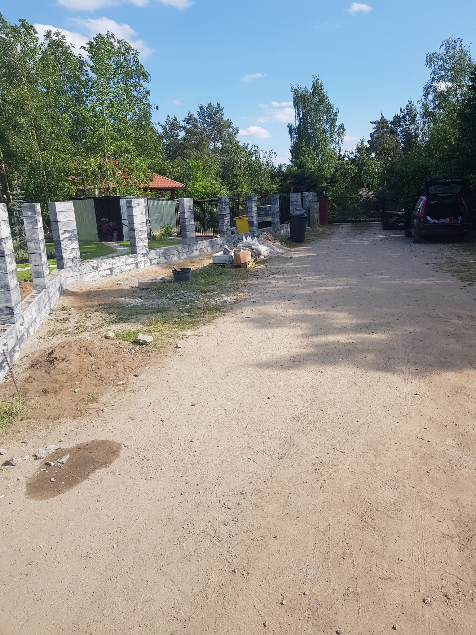 Budowa ogrodzenia z prefabrykowanych elementów betonowych, widoczne słupki i podmurówka w trakcie montażu, teren budowy z workami cementu i narzędziami, samochód osobowy w tle.