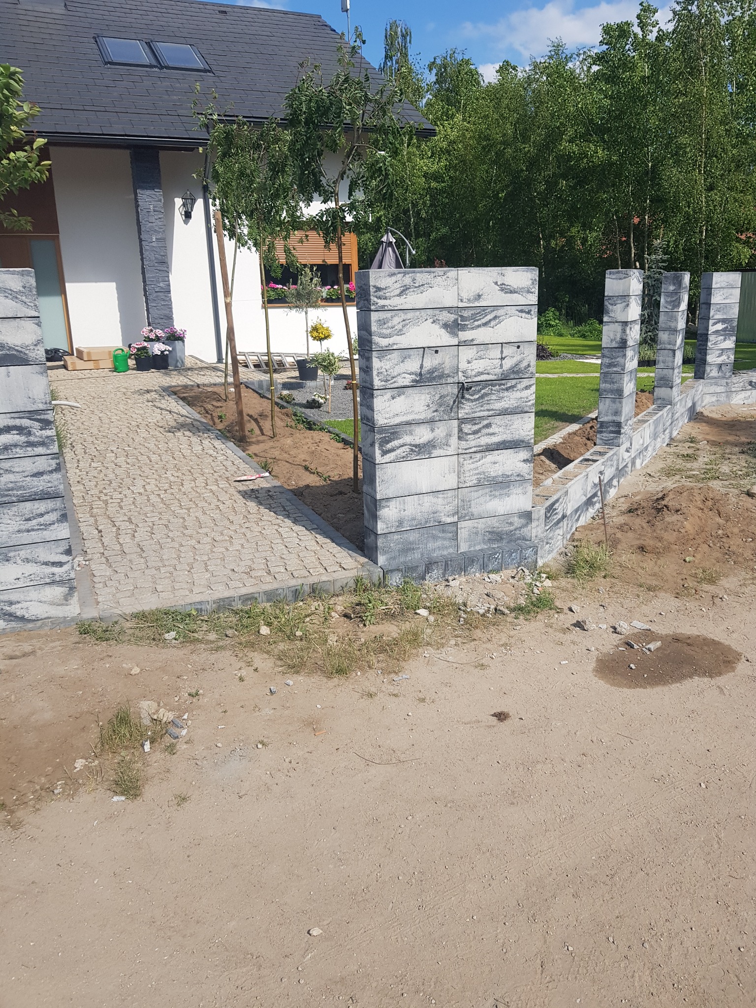 Budowa ogrodzenia z szarych, betonowych bloczków o marmurkowym wzorze, widoczny fragment posesji z kostką brukową i dom w tle.