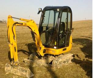 Żółta mini koparka gąsienicowa JCB na nierównym, ziemistym terenie. Widoczna kabina operatora i ramię koparki z łyżką. Tło stanowią pagórki i zabudowania.