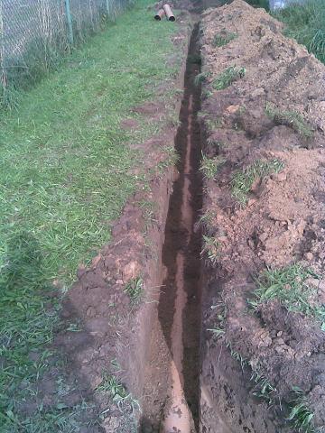 Wykopany wąski rów w ziemi z widoczną rurą na dnie, obok rów z pryzmą ziemi i zielona trawa.