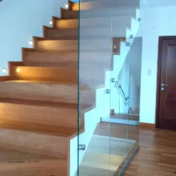 Drewniane schody z oświetleniem punktowym na stopniach i szklaną balustradą, widoczne drewniane podłogi i drzwi.