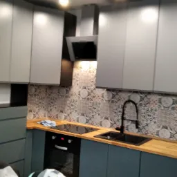 Szare szafki kuchenne z drewnianym blatem, czarnym zlewem i baterią na tle dekoracyjnych płytek ściennych.